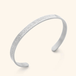 A silver, open-cuff bangle bracelet with an intricate, repeating pattern of Arabic script around its entire outer surface. The bracelet is displayed on a light beige background.