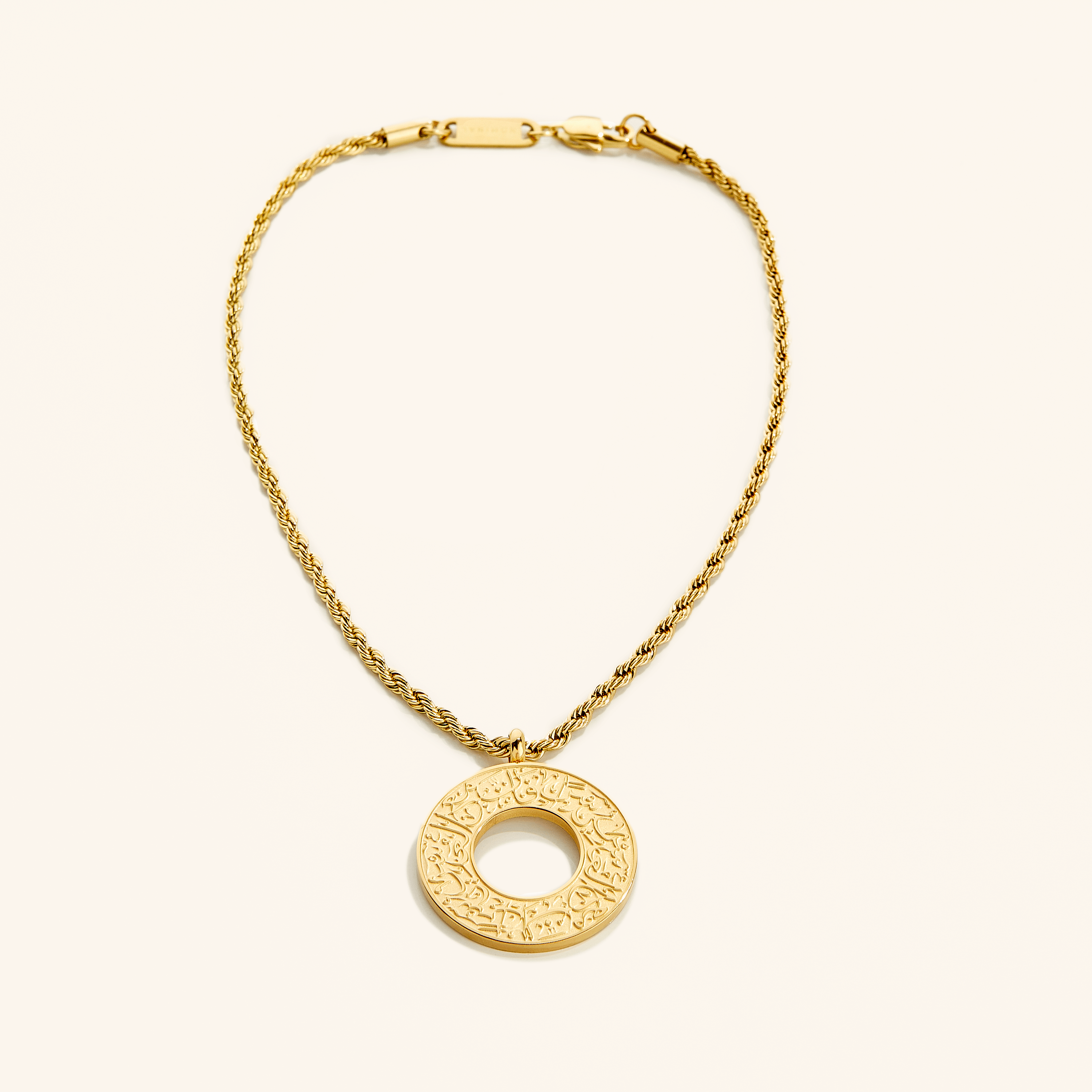 Gold car hanger with a circular pendant engraved in Arabic calligraphy reading “With Hardship Comes Ease,” displayed against a cream background.