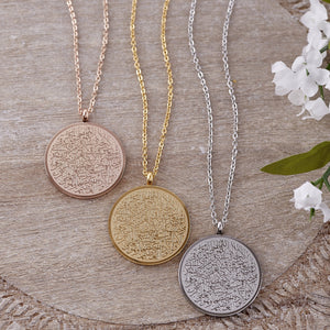 Three necklaces with round pendants intricately inscribed with Arabic script are displayed on a textured wooden surface. From left to right, the necklaces are rose gold, gold, and silver, each with a delicate chain. White flowers with green leaves are visible in the upper right corner of the image, adding a soft, natural element to the composition. Given the inscription and the product name "Ayatul Kursi Necklace," it is highly likely that the script is from the Quran, a central religious text in Islam.