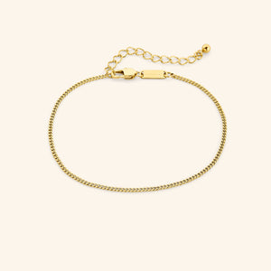 Gold curb chain anklet with a lobster clasp and extender, featuring a tag engraved with "NOMINAL" near the clasp, laid out on a neutral background.