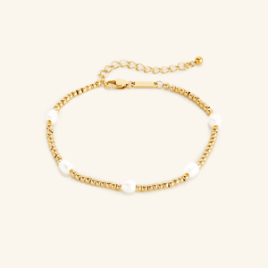 Gold anklet with small round beads and five evenly spaced white pearls, shown on a cream background with an adjustable clasp and extender chain engraved with 'NOMINAL'.