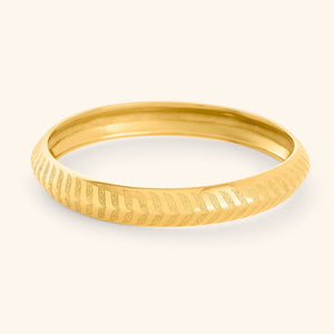 A gold "Chevron Bangle" designed for women as part of the Aadil x Nominal collection by Nominal. 1 The bangle has a smooth, rounded surface and is decorated with intricate, stylized chevron or feather-like carvings that run around its entire circumference