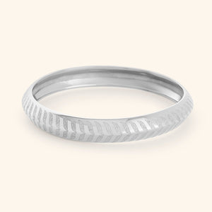 A silver "Chevron Bangle" designed for women as part of the Aadil x Nominal collection by Nominal. The bangle has a smooth, rounded surface and is decorated with intricate, stylized chevron or feather-like carvings that run around its entire circumference.
