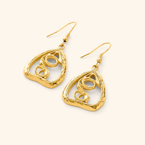 A pair of gold "Hub (Love)" earrings. The earrings feature a unique, abstract design with swirling lines enclosed within a roughly triangular frame. The metal is a warm gold color, and the background is a soft, light beige. The image is a product shot, likely showcasing the design and craftsmanship of the earrings. The text "This image might show: Hub (Love) Earrings" confirms the product name.