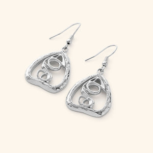 A pair of silver "Hub (Love)" earrings. The earrings feature a unique, abstract design with swirling lines enclosed within a roughly triangular frame. The metal is a warm gold color, and the background is a soft, light beige. The image is a product shot, likely showcasing the design and craftsmanship of the earrings. The text "This image might show: Hub (Love) Earrings" confirms the product name.