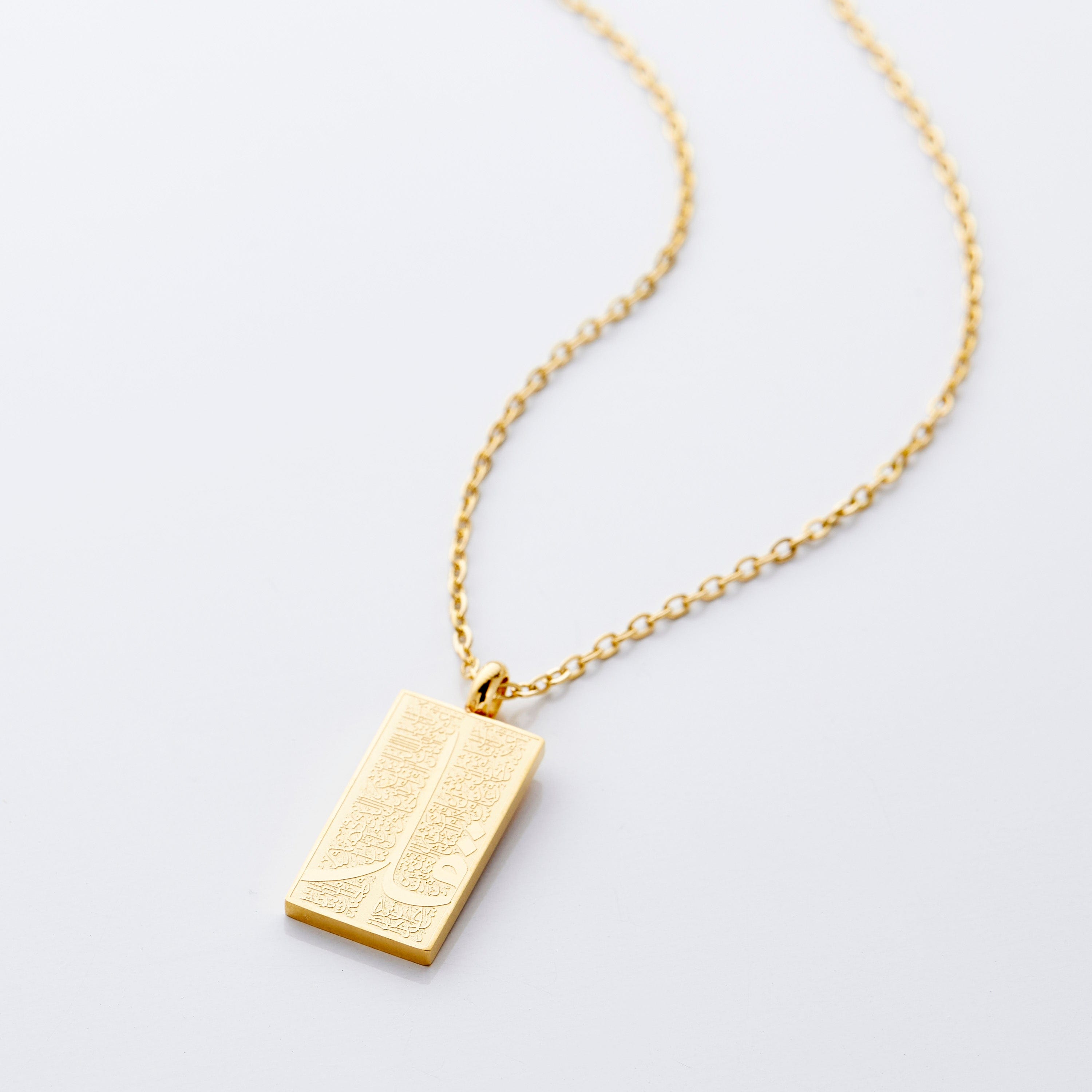 The image shows a delicate gold necklace displayed against a clean, light gray background. The chain is thin and elegant, forming a gentle curve that draws the eye toward the pendant hanging at the center. The pendant is rectangular and made of gold, with a smooth, polished finish that reflects light subtly. Its surface features intricate engraved patterns that add texture and visual interest.