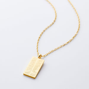 The image shows a delicate gold necklace displayed against a clean, light gray background. The chain is thin and elegant, forming a gentle curve that draws the eye toward the pendant hanging at the center. The pendant is rectangular and made of gold, with a smooth, polished finish that reflects light subtly. Its surface features intricate engraved patterns that add texture and visual interest.