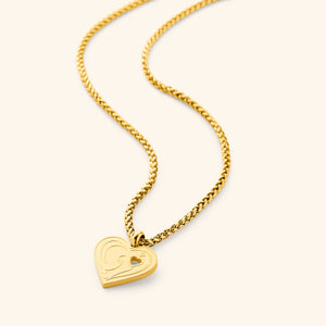 A close-up of the "Piece of Me" Mama Necklace in 18K gold plating, featuring a heart-shaped pendant engraved with the Arabic word "Ummi" ("my mother") in elegant calligraphy. A small heart-shaped cutout is present on the pendant, symbolizing a piece given to a loved one. The pendant hangs from a delicate wheat chain.