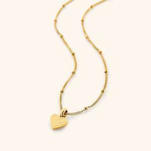 A close-up of the Mother Love Necklace in 18K gold plating, featuring a minimal heart-shaped charm engraved with the Arabic word "Ummi" ("my mother") in elegant calligraphy. The pendant hangs from a delicate dotted chain, showcasing its minimalist design.
