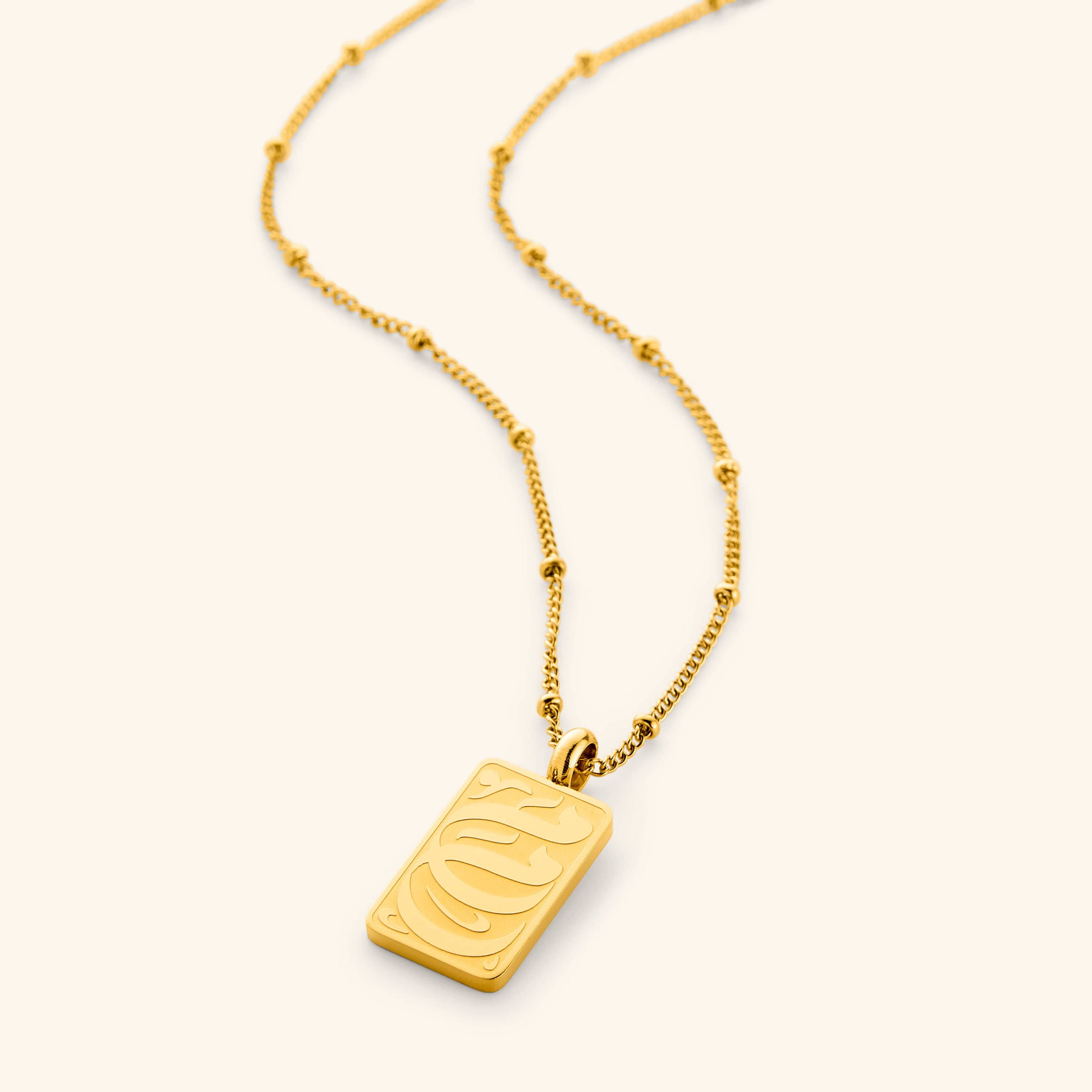 A close-up of the "My Mother Tag Necklace" in 18K gold plating, featuring a rectangular pendant engraved with the Arabic word "Ummi" ("my mother") in elegant calligraphy. The pendant hangs from a delicate dotted chain, showcasing its minimalist design.