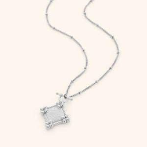 Photo of a necklace with silver diamond-shaped plaque stamped with حَسْبُنَا اللَّهُ وَنِعْمَ الْوَكِيلُ (Hasbunallahu wa ni'mal wakeel). "Allah is sufficient for us, and He is the best Disposer of affairs." framed in beaded borders on a thin dotted chain.