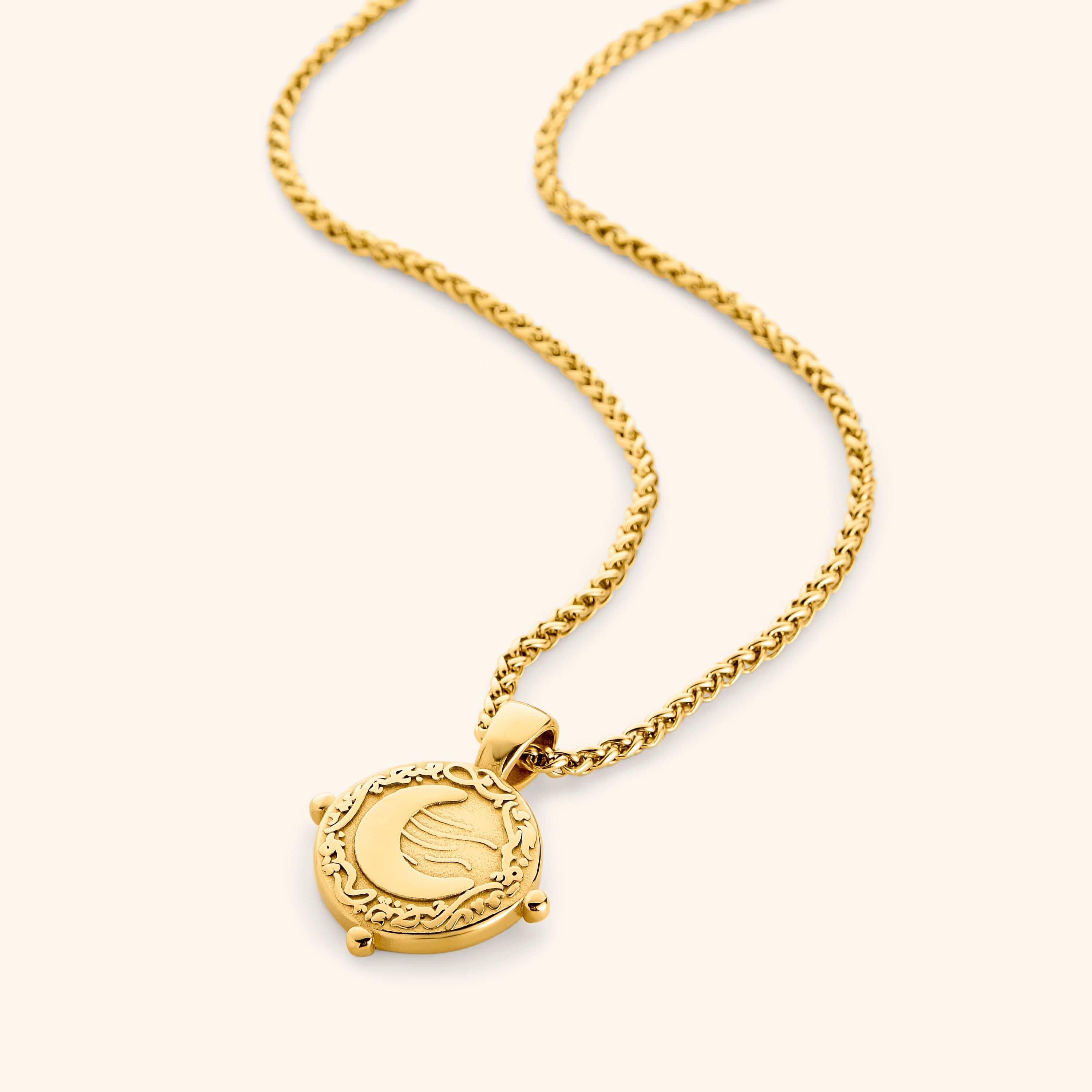Gold necklace with a round pendant featuring the engraved phrase 'What you seek is seeking you' in Arabic calligraphy. The design includes a raised crescent moon and a flowing motif, framed with vine detailing and three small bead accents around the border.