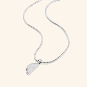 A silver necklace with a pendant shaped like a slice of a watermelon. The watermelon pendant has small indentations representing seeds. The necklace features a thin, flexible silver snake chain. The necklace is displayed on a light beige background.
