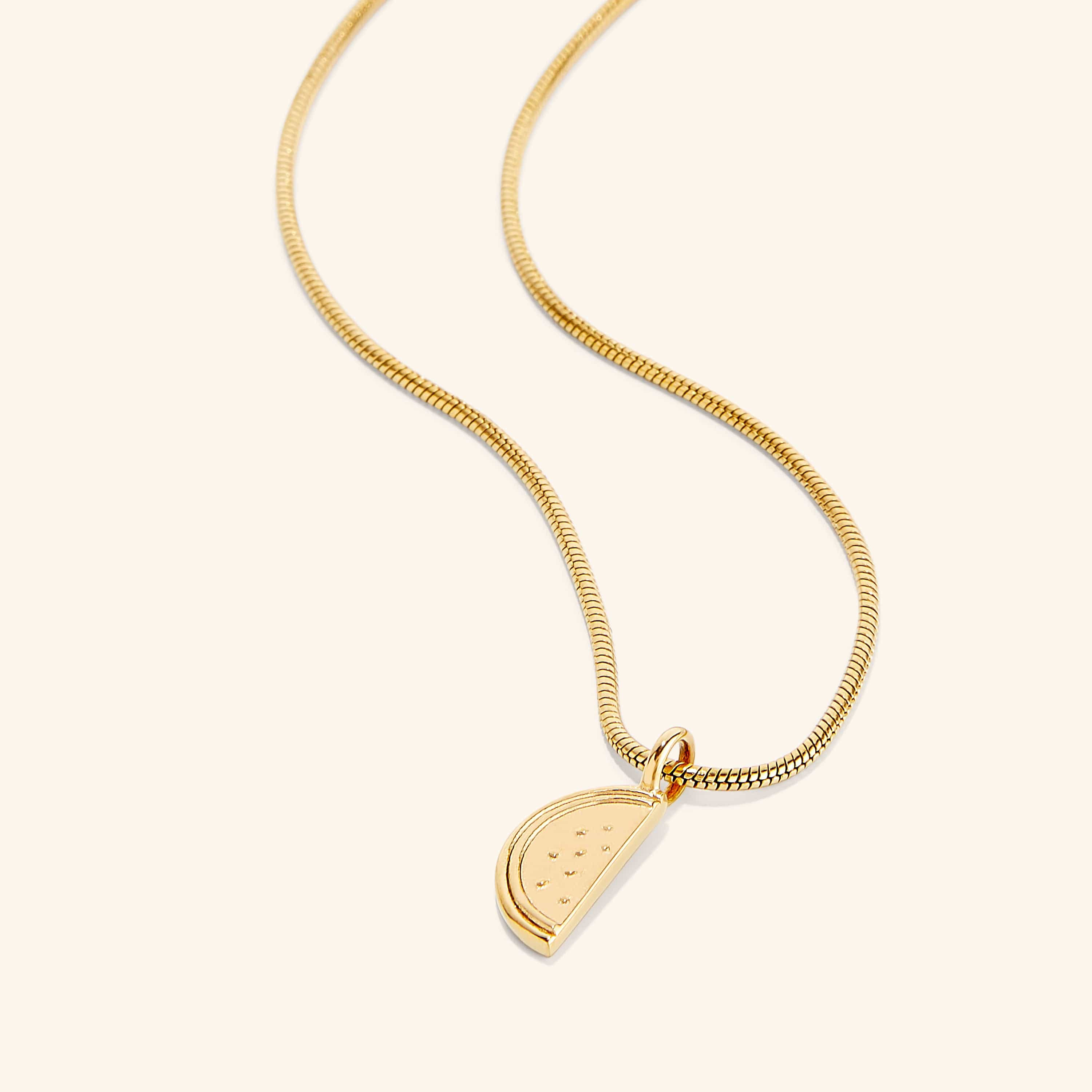 A gold necklace with a pendant shaped like a slice of a watermelon. The watermelon pendant is detailed with small indentations representing seeds. The necklace chain is a thin, flexible gold snake chain. The necklace is displayed against a light beige background.