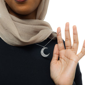 A close-up of a silver necklace with a crescent moon pendant. The pendant is inscribed with Arabic script. The necklace is held up against a blurred background of a person wearing a black top. The focus is on the detail of the pendant and the script. Given the inscription "With Hardship Comes Ease" and the crescent moon, which holds significance in Islamic culture, it's plausible that the script is related to Islamic teachings and the necklace carries religious or inspirational meaning for the wearer.