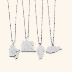 Four silver "With Hardship Comes Ease" Map Necklaces arranged side by side on a white background, each pendant shaped like a different map and engraved with Arabic calligraphy, showcasing subtle differences in map outlines and silver chain styles.