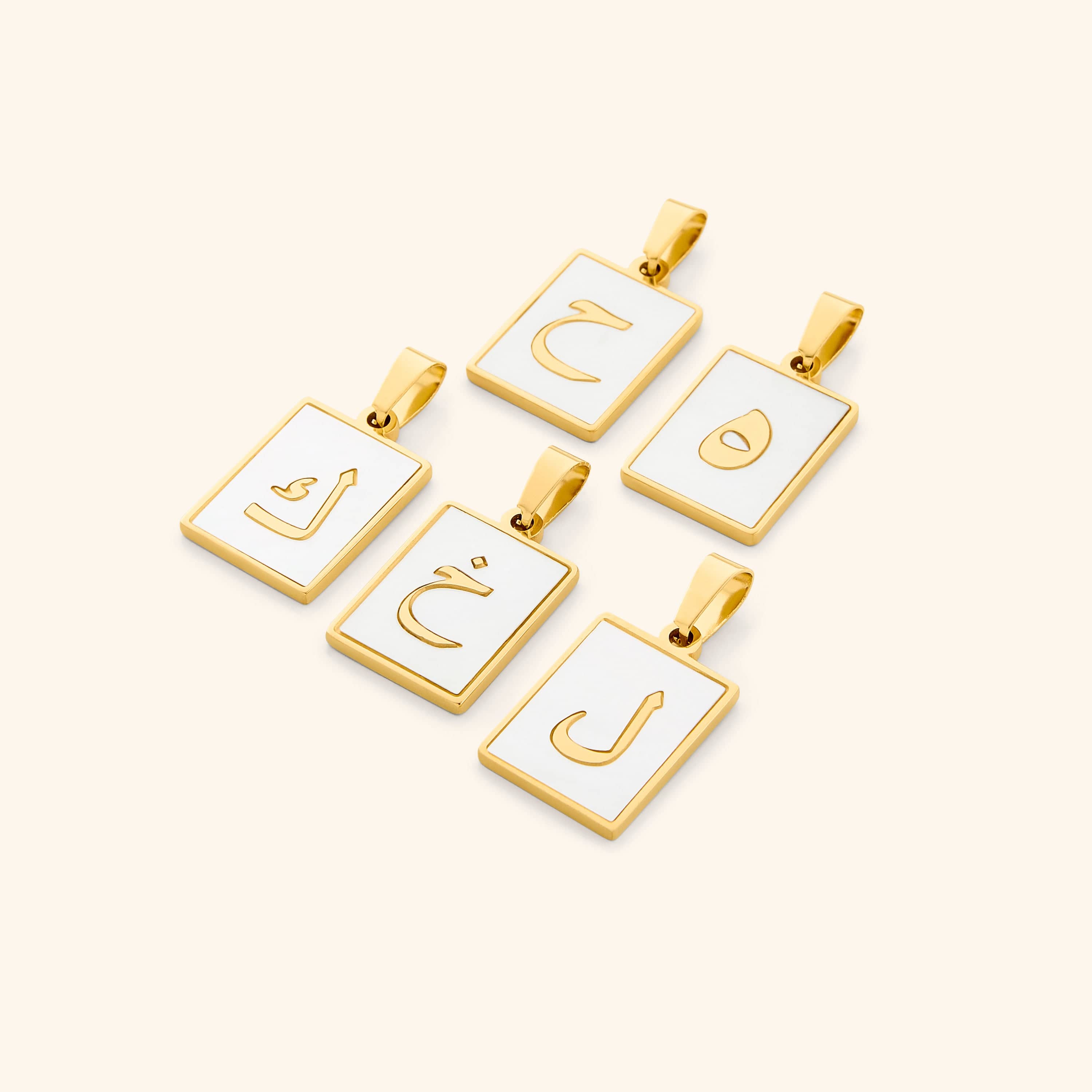 A flat lay of five Arabic letter charms in 18K gold plating, each featuring an iridescent pearl backdrop. The charms are engraved with Arabic letters.