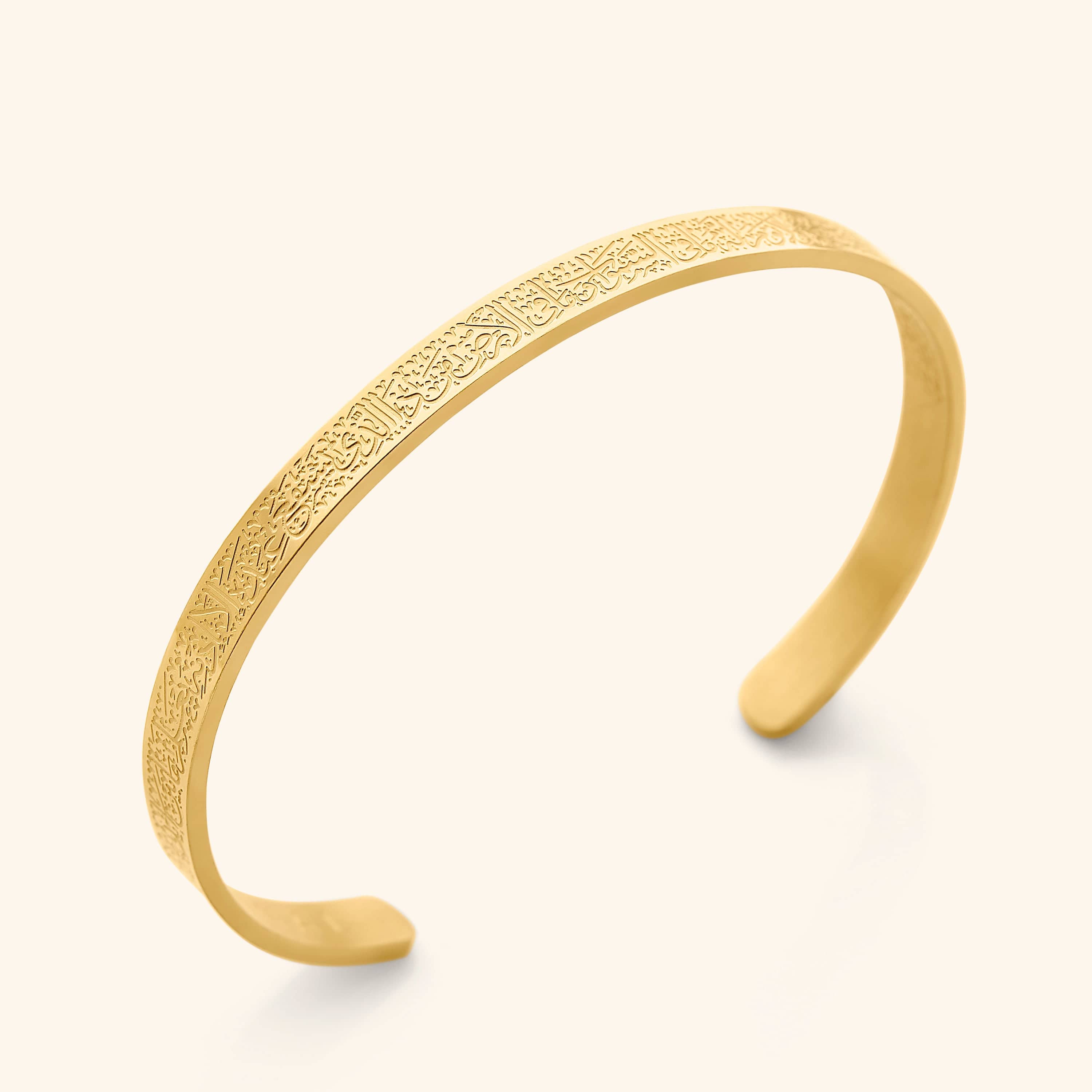 A close-up shot showcases a slender, open-ended gold cuff bracelet. The bracelet's surface is intricately engraved with Arabic script, identified as the Ayatul Kursi. The bracelet has a smooth, polished finish and rests on a soft, off-white surface, casting a subtle shadow beneath.