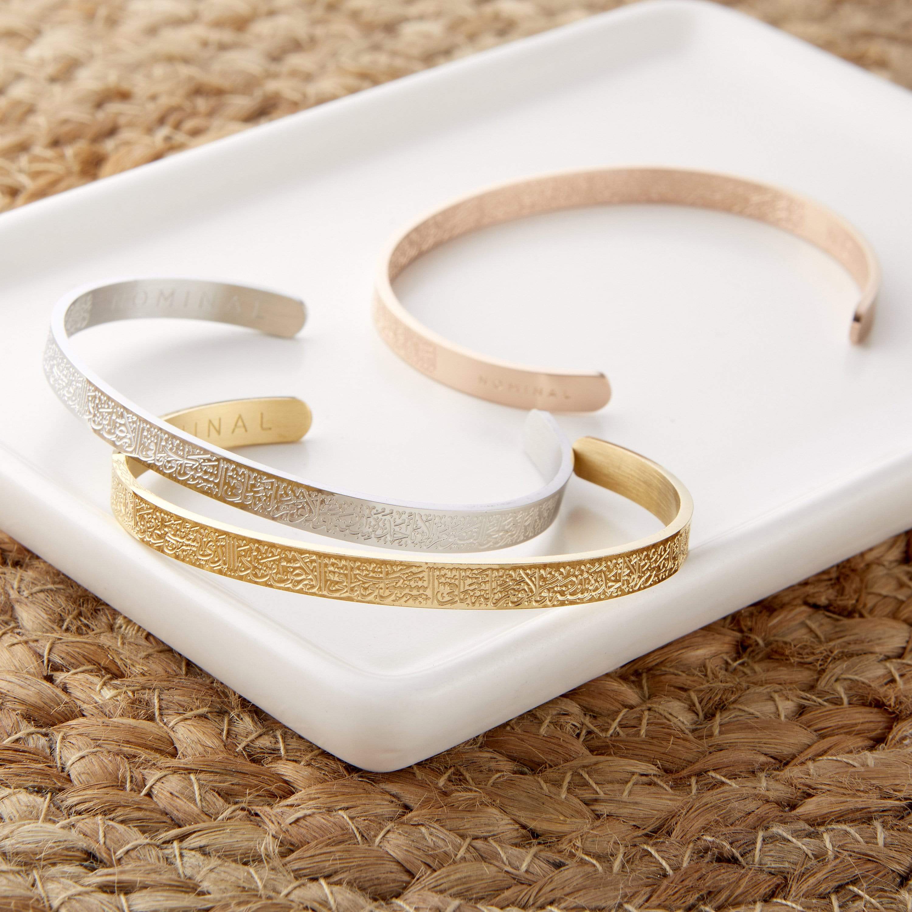Three open-cuff bangle bracelets are arranged on a white rectangular tray. The bracelet at the top is rose gold and appears smooth. The bracelet in the middle is silver and has an intricate pattern of Arabic script etched around it, with the word "NOMINAL" visible on the inner side. The bracelet at the bottom is gold and also features an intricate pattern of Arabic script. The tray rests on a textured, woven surface, possibly a placemat or table covering.