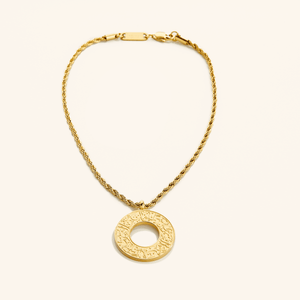 Gold car hanger with a circular pendant engraved in Arabic calligraphy reading “With Hardship Comes Ease,” displayed against a cream background.