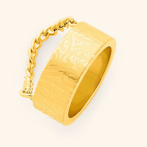 A wide, gold-plated ring with a rectangular face and intricate, abstract designs engraved across the surface. A gold chain is attached to either side of the ring. The ring is displayed against a light beige background.