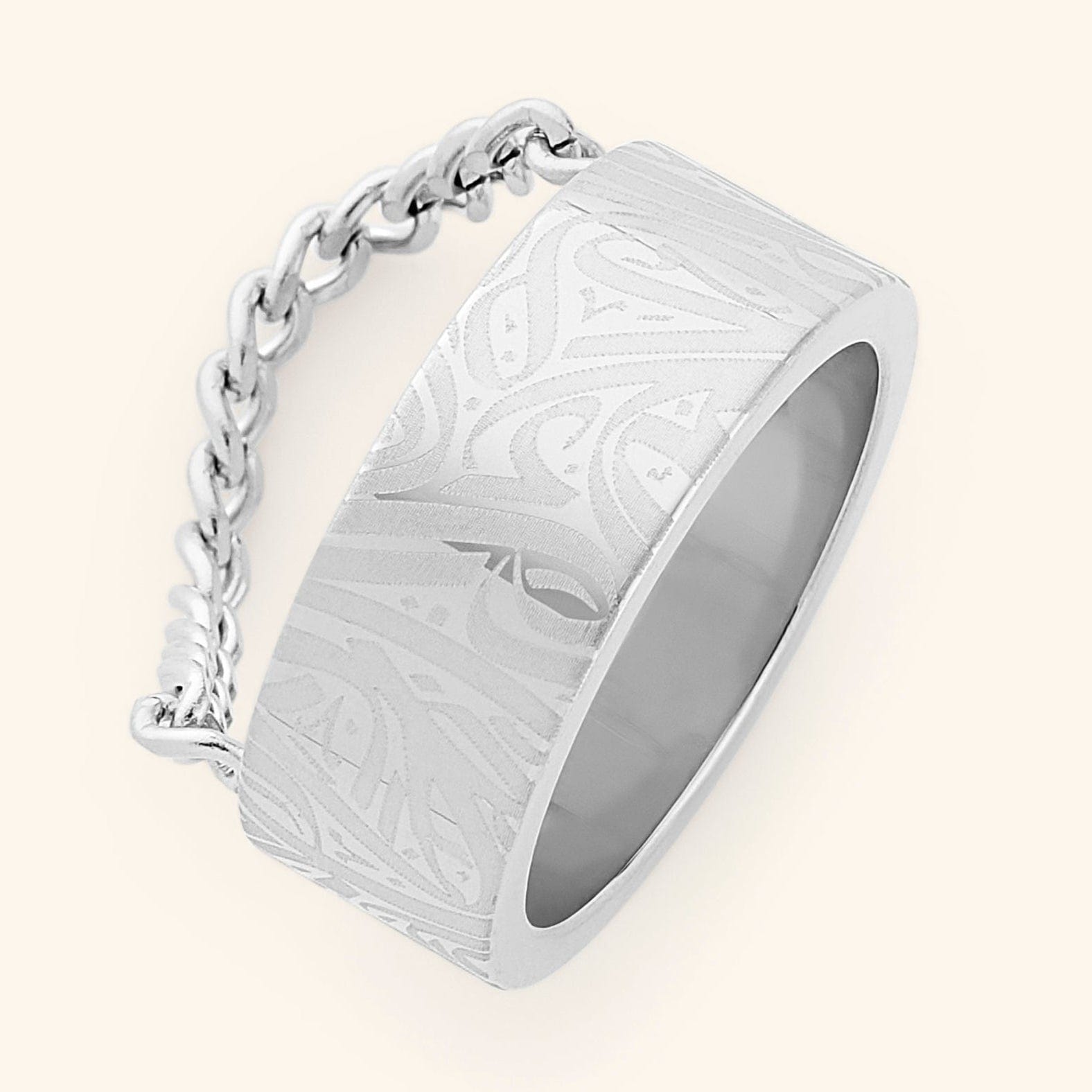 A wide, silver-plated ring with a rectangular face and intricate, abstract designs engraved across the surface. A delicate silver chain is attached to either side of the ring. The ring is displayed against a light beige background.