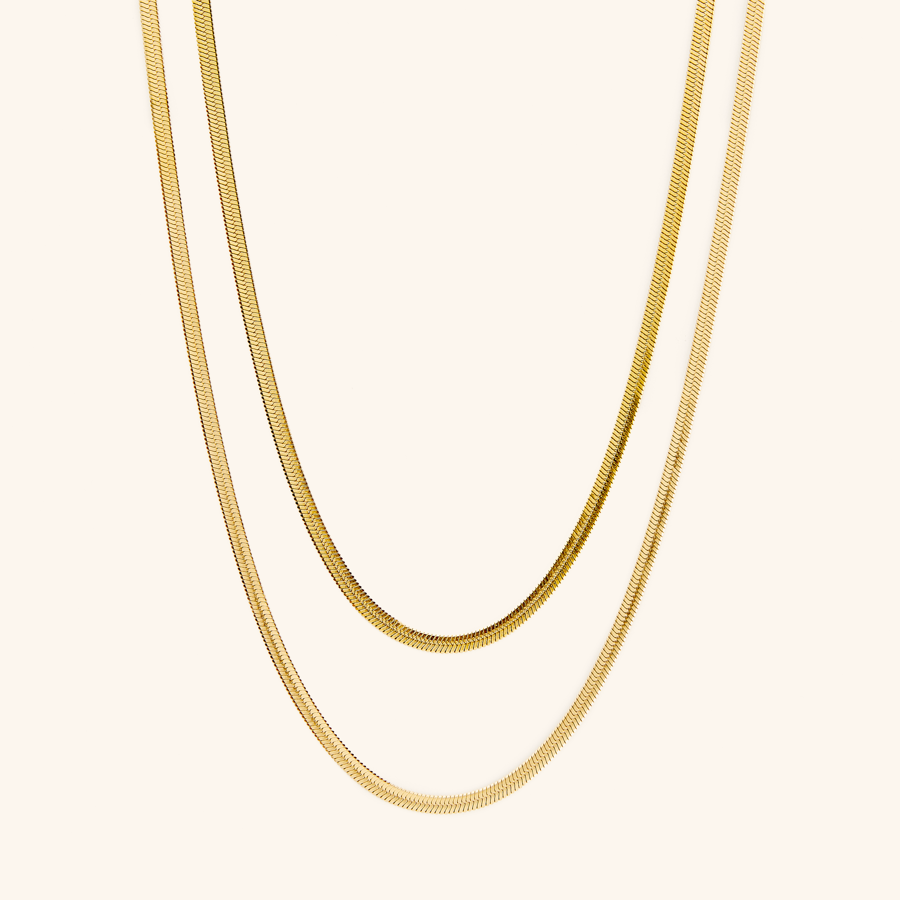 Two gold herringbone chains laid flat against a cream background.