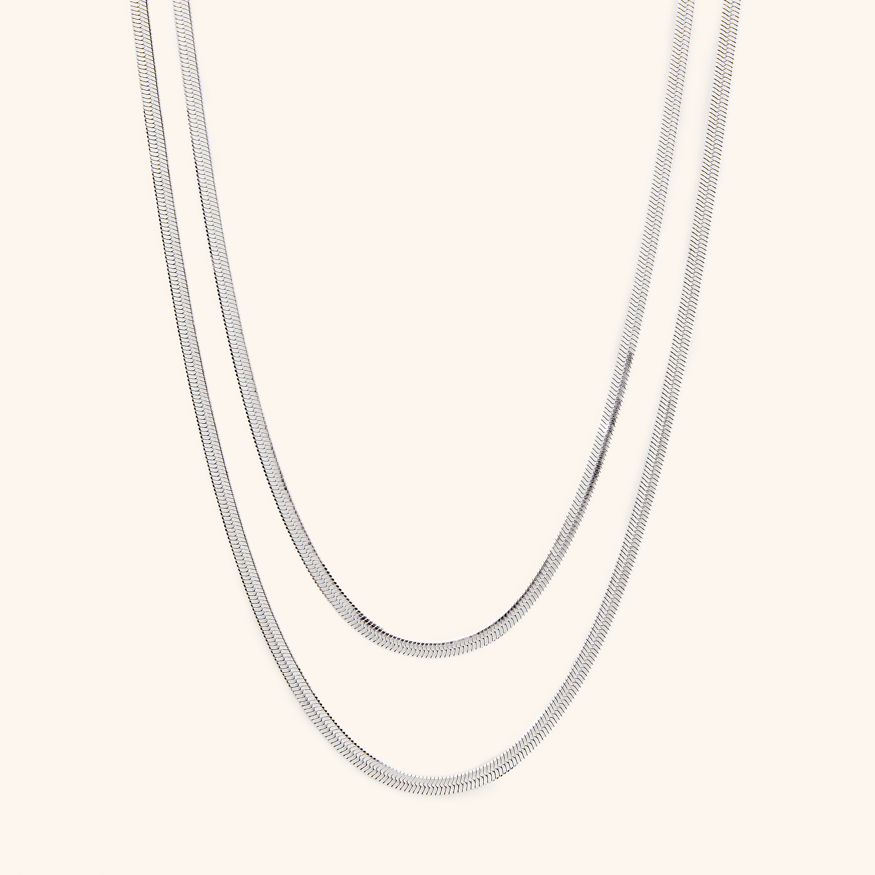  Two silver herringbone chains laid flat against a cream background.