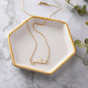 A gold necklace with a name pendant in Arabic script. The chain is delicate and gold. The necklace is displayed on a light beige background.