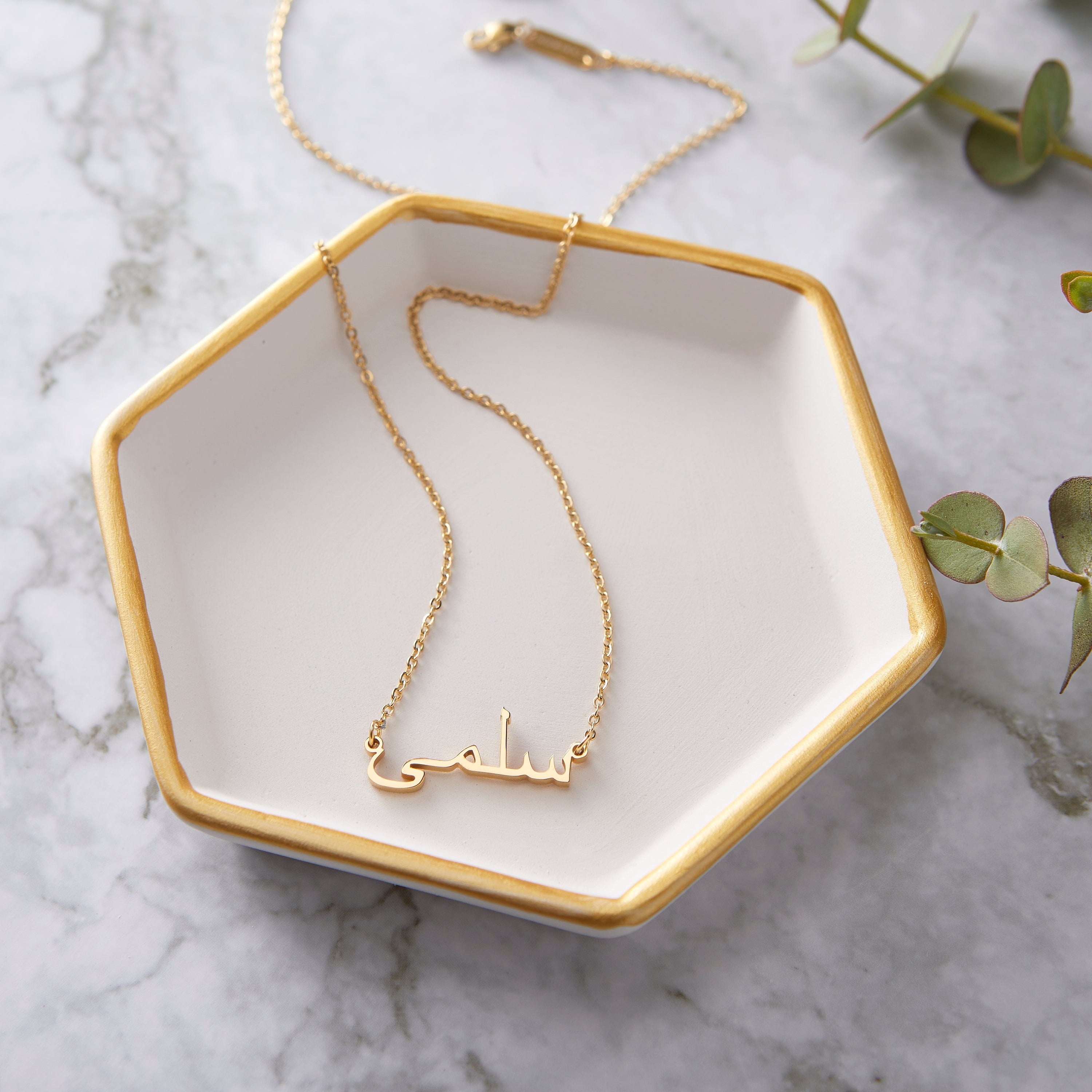 A gold necklace with a name pendant in Arabic script. The chain is delicate and gold. The necklace is displayed on a light beige background.