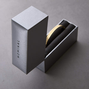 Silver Nominal drawer-style gift box with black text “NOMINAL” on the side, opened vertically to reveal a gold cuff bracelet resting in black foam lining, styled on a gray background.