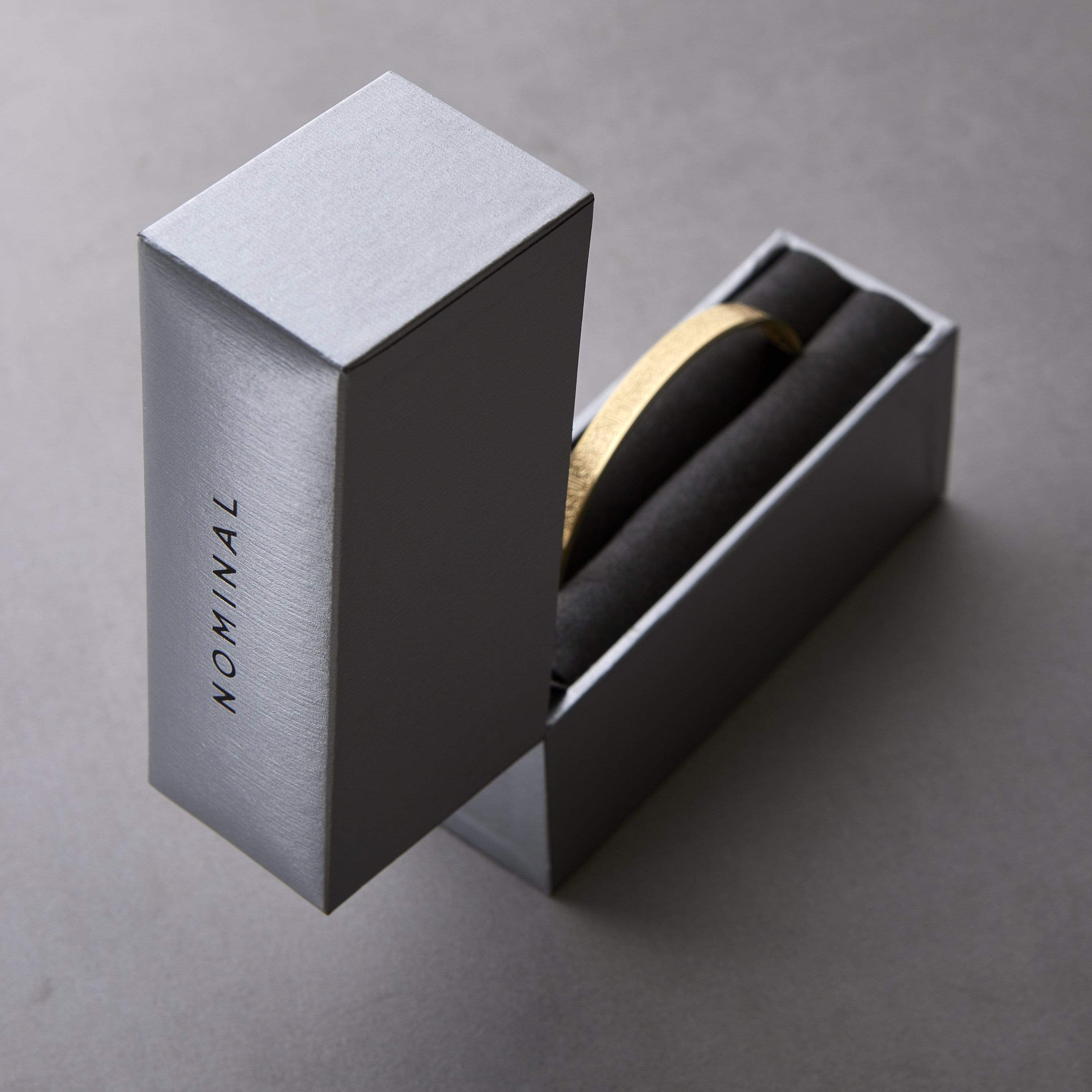 Silver Nominal drawer-style gift box with black text “NOMINAL” on the side, opened vertically to reveal a gold cuff bracelet resting in black foam lining, styled on a gray background.