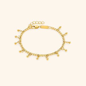 Flat lay of a gold dangling dotted bracelet featuring small dangling beads, displayed against a cream background. The clasp tag shows the engraved text “NOMINAL”.