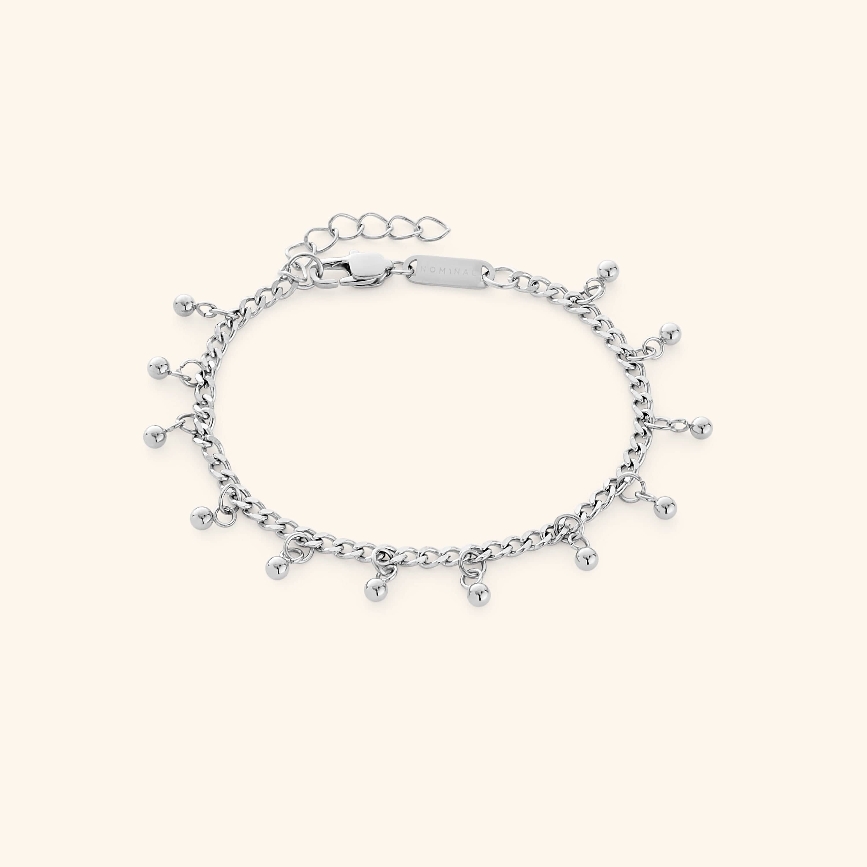 Flat lay of a silver dangling dotted bracelet featuring small dangling beads, displayed against a cream background. The clasp tag shows the engraved text “NOMINAL”.