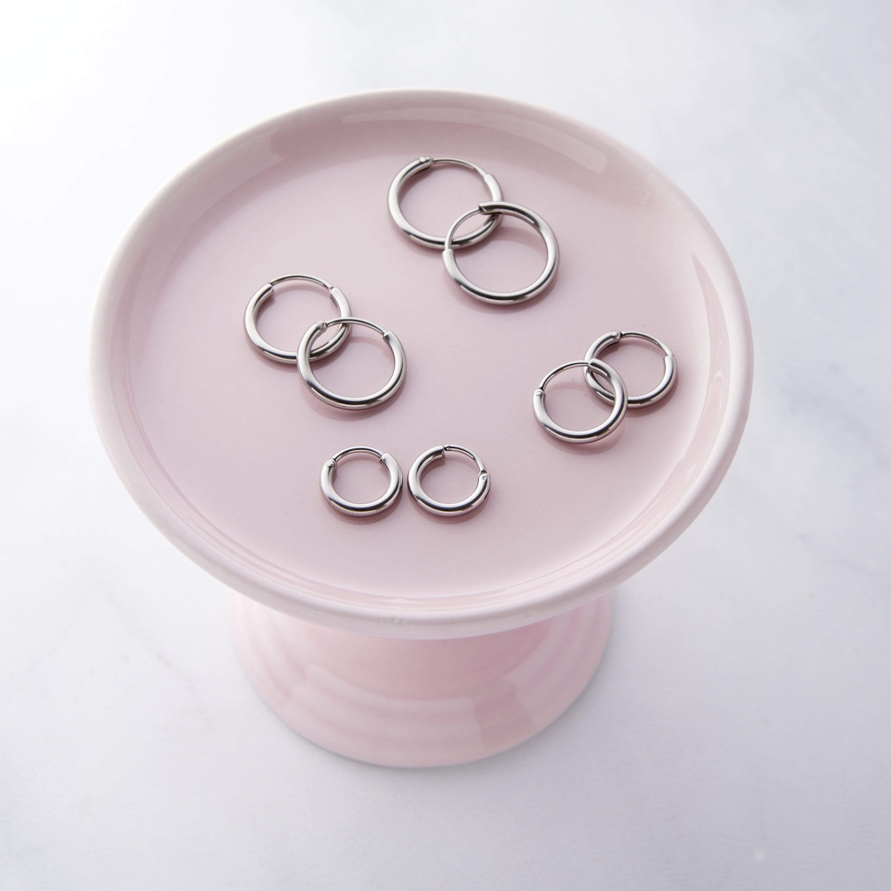 Flat lay of four pairs of polished silver hoop earrings in varying sizes, arranged on a pale pink pedestal dish.