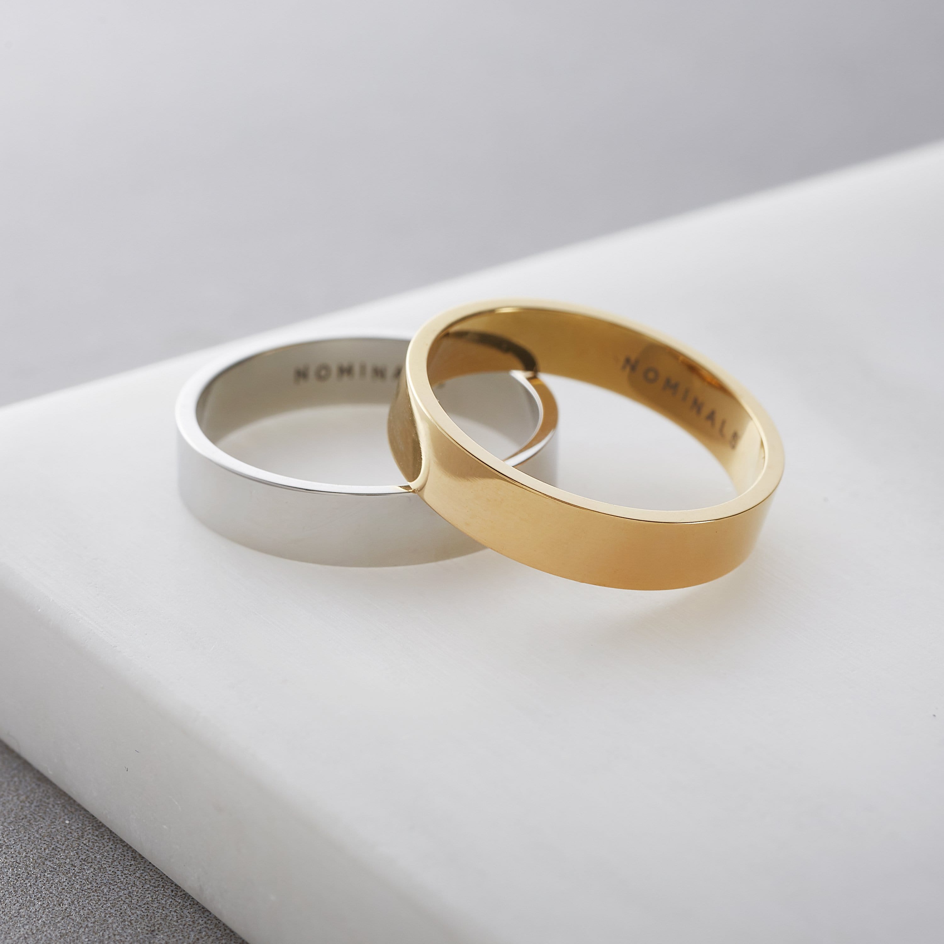 Gold and silver band rings resting together on a white marble surface, engraved with 'NOMINAL' on the inside.