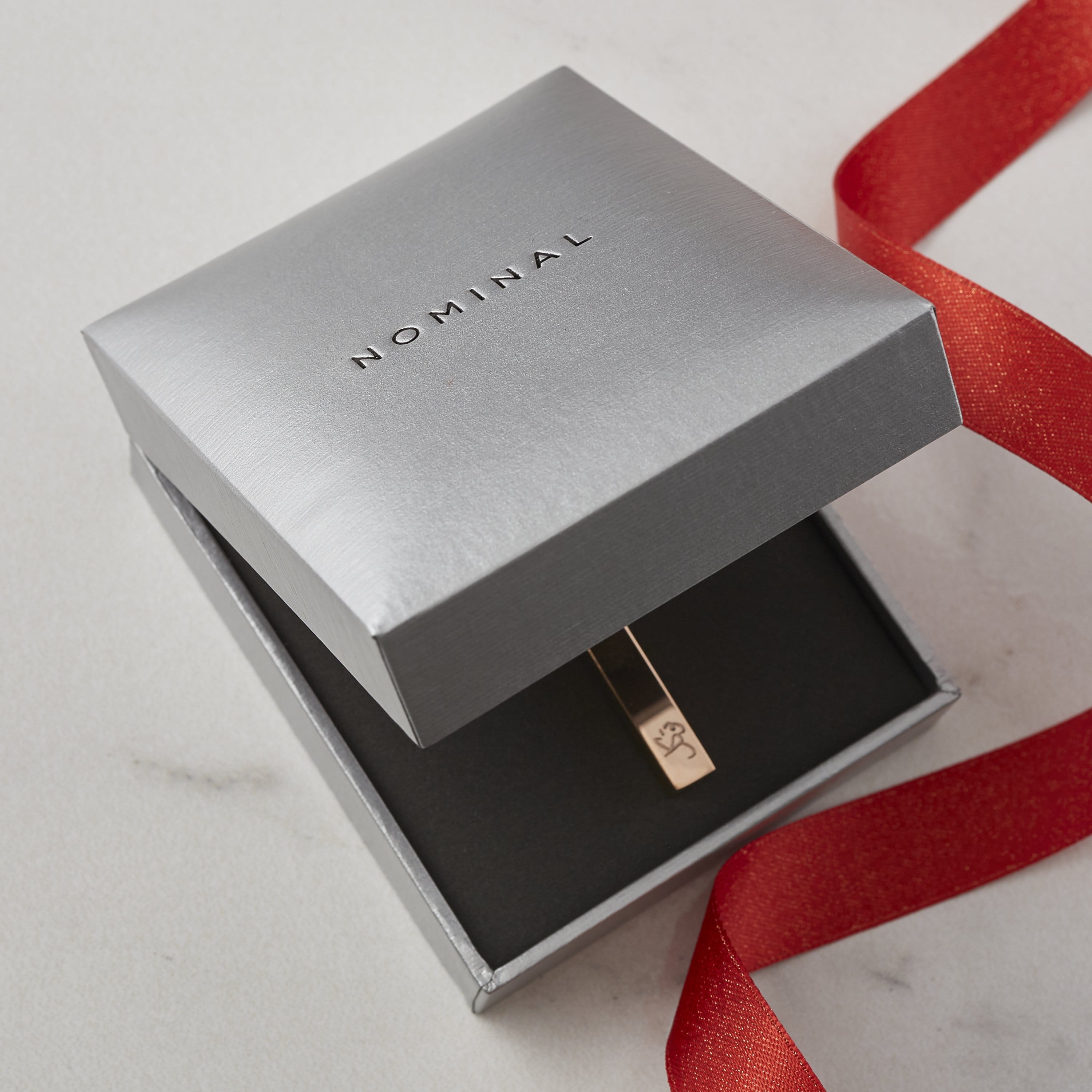 Silver Nominal gift box with black text “NOMINAL” on the lid, partially open to reveal a gold bar necklace inside, styled with a red ribbon on a light surface.