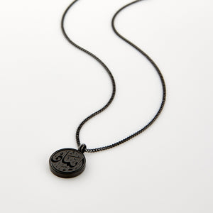 A black Faith Token necklace laid flat on a white background, showing the engraved Arabic script “Iman”.