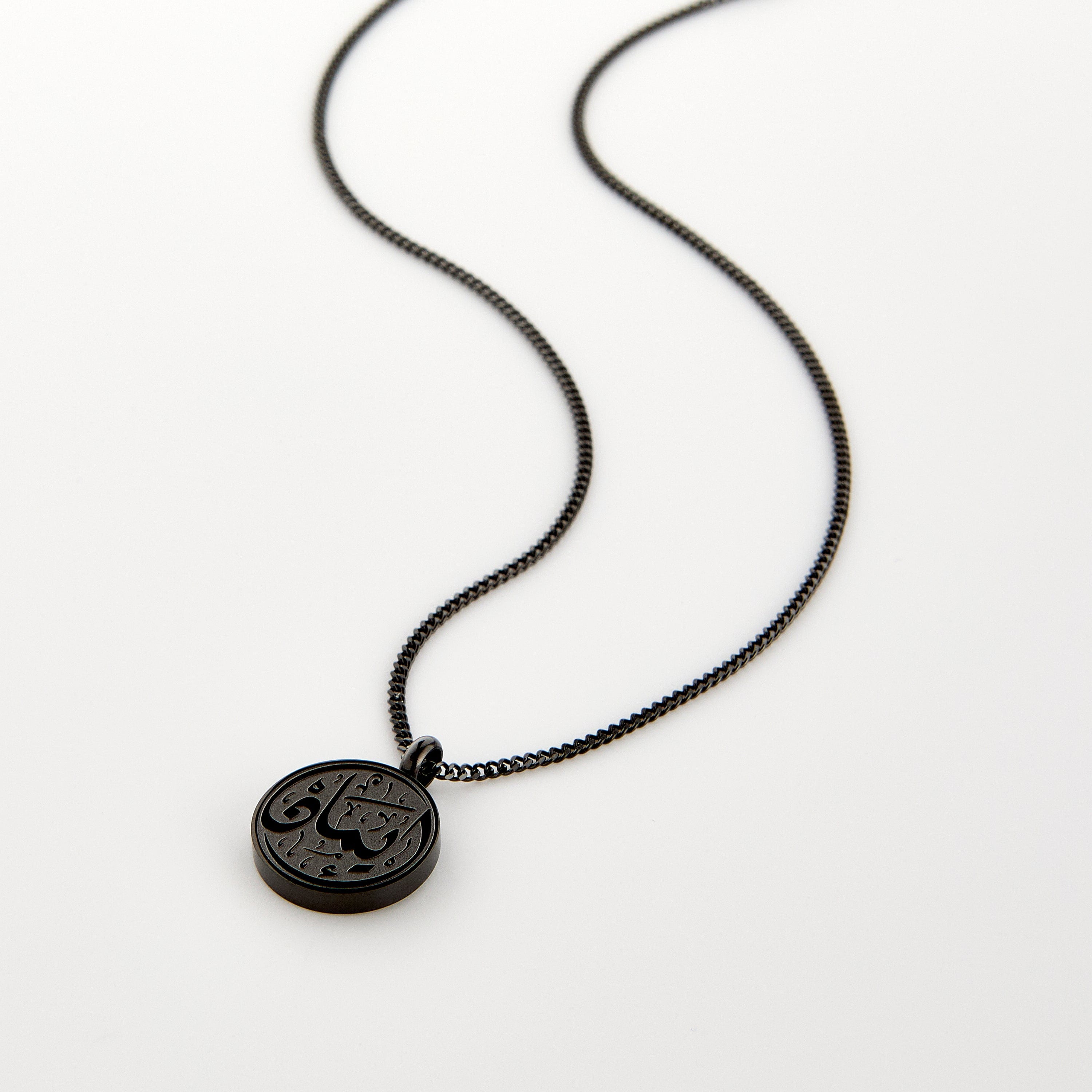 A black Faith Token necklace laid flat on a white background, showing the engraved Arabic script “Iman”.