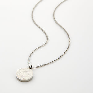 A silver Faith Token necklace with the pendant resting flat on a light background, showing the engraved Arabic script “Iman”.