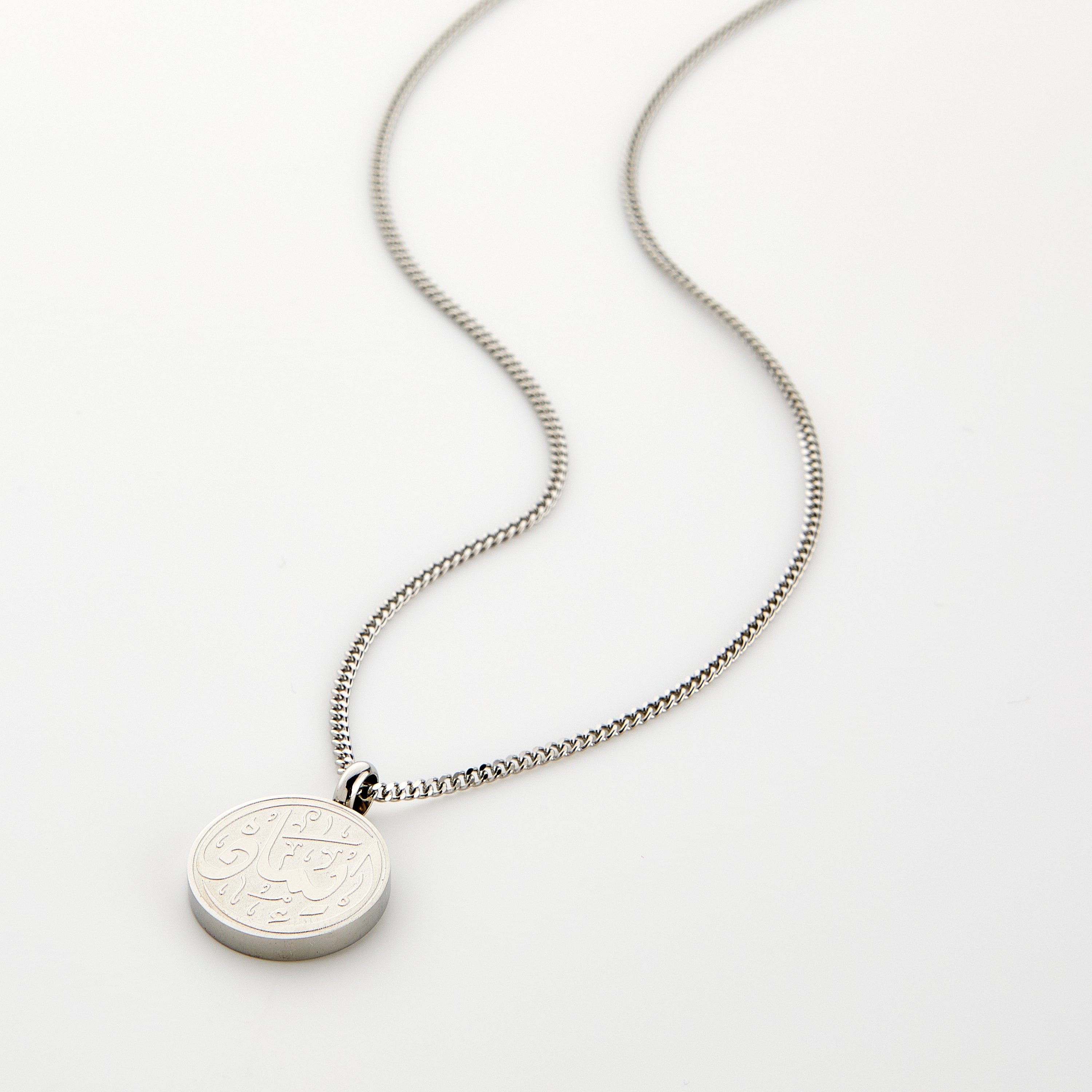 A silver Faith Token necklace with the pendant resting flat on a light background, showing the engraved Arabic script “Iman”.
