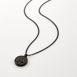 Black necklace with a circular pendant engraved with 'Tawakkul' calligraphy, displayed on a white background.