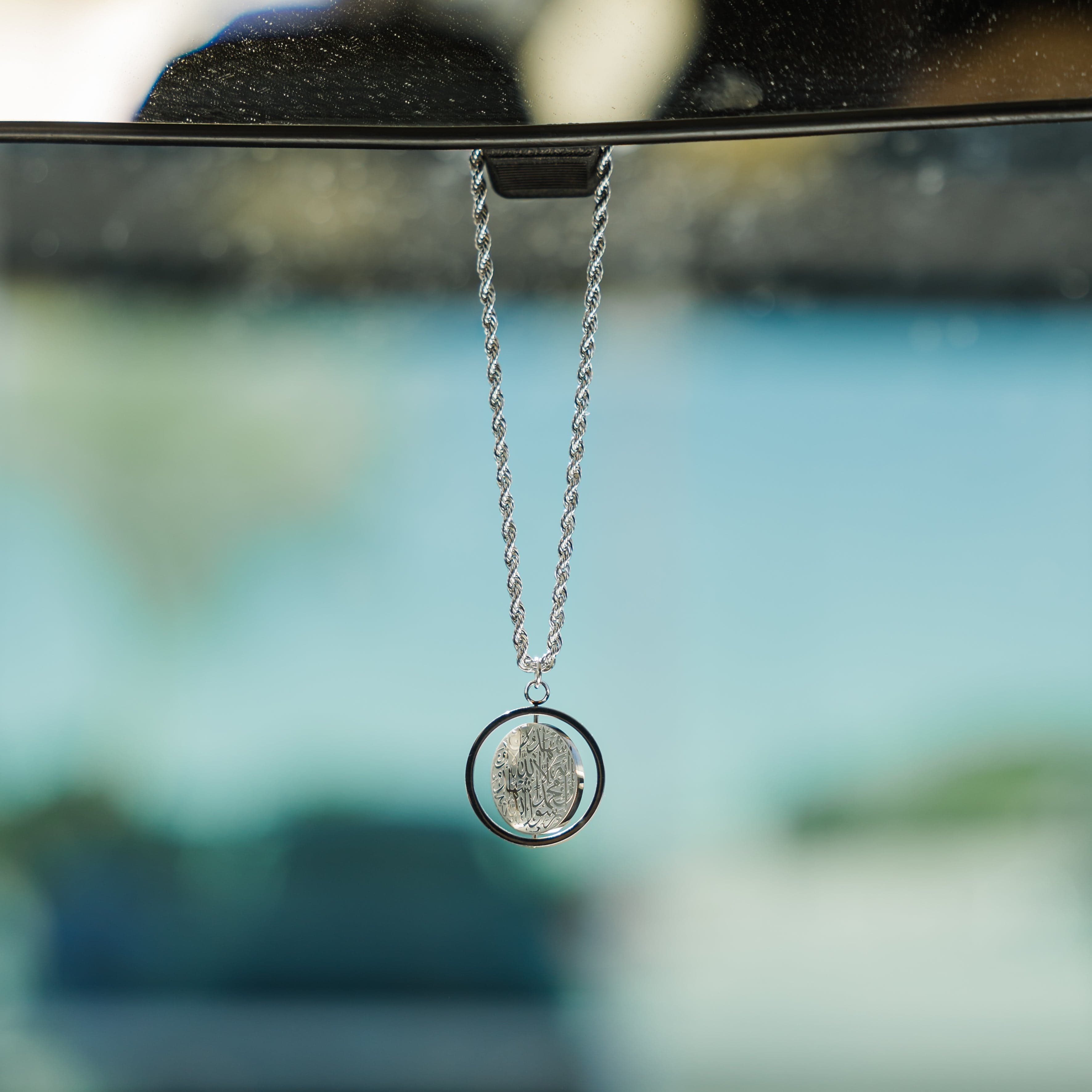 Close-up of a silver car hanger with a circular pendant engraved in Arabic calligraphy of the Shahada.