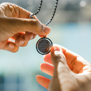 Hands holding a black car hanger with a circular pendant engraved in Arabic calligraphy of the Shahada, photographed against a bright background.