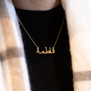 A close-up shows a hand gently holding a gold necklace with a pendant featuring a name written in elegant Arabic script. The necklace has a delicate gold chain. The background includes a black ribbed top and a portion of a rust-colored garment draped over the shoulder. The focus is on the intricate detail of the Arabic name pendant and the delicate nature of the jewelry.