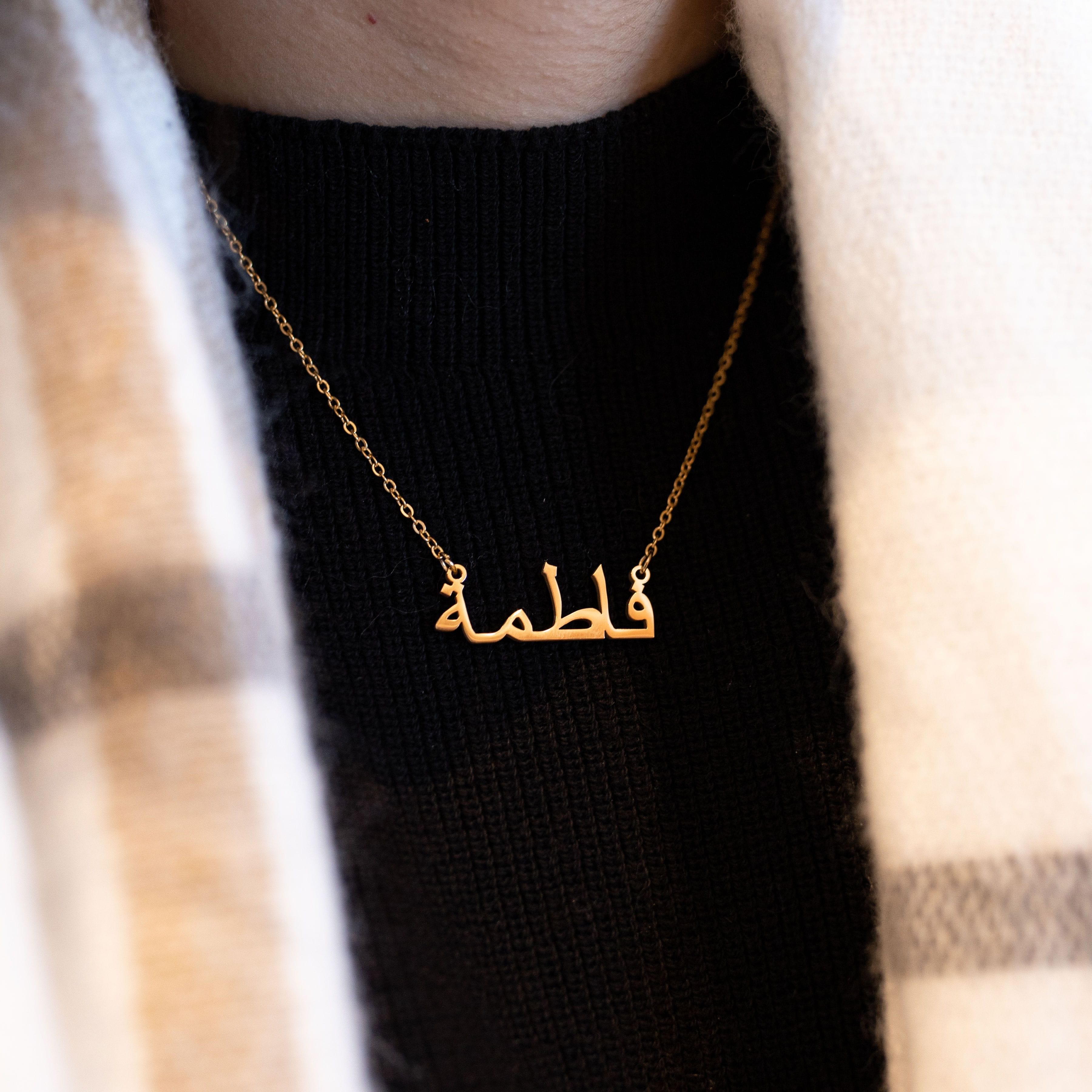 A close-up shows a hand gently holding a gold necklace with a pendant featuring a name written in elegant Arabic script. The necklace has a delicate gold chain. The background includes a black ribbed top and a portion of a rust-colored garment draped over the shoulder. The focus is on the intricate detail of the Arabic name pendant and the delicate nature of the jewelry.