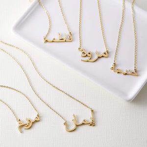 Five gold necklaces with pendants featuring different names written in Arabic script are arranged on a white rectangular tray. Each necklace has a delicate gold chain. The names on the pendants are distinct in their calligraphic style. The tray rests on a textured, light-colored surface.