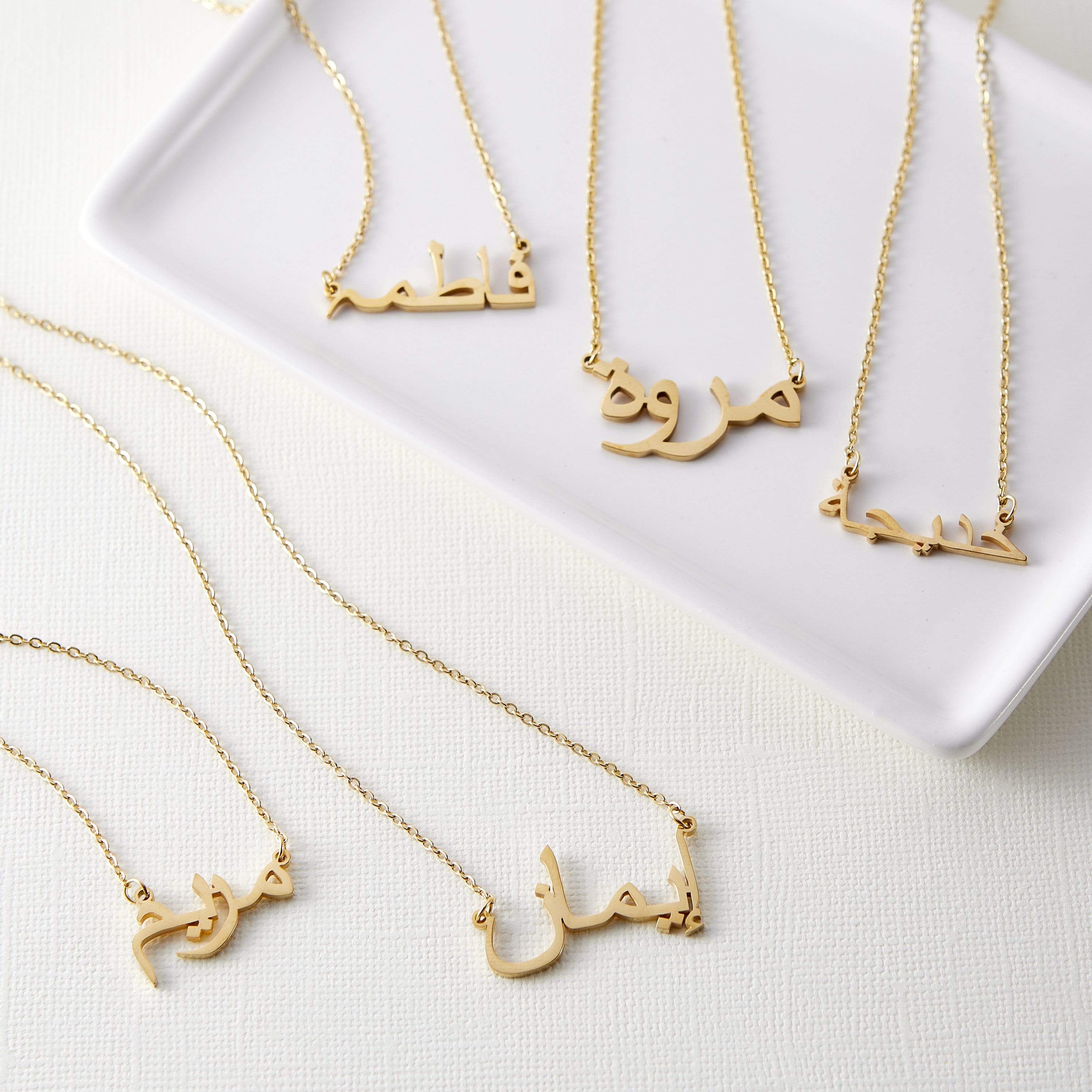 Five gold necklaces with pendants featuring different names written in Arabic script are arranged on a white rectangular tray. Each necklace has a delicate gold chain. The names on the pendants are distinct in their calligraphic style. The tray rests on a textured, light-colored surface.