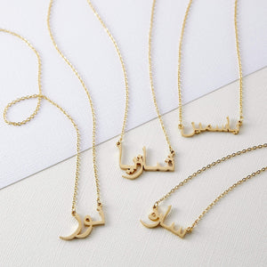 Four gold necklaces with pendants featuring different names written in Arabic script are arranged on a white rectangular tray. Each necklace has a delicate gold chain. The names on the pendants are distinct in their calligraphic style. The tray rests on a textured, light-colored surface.
