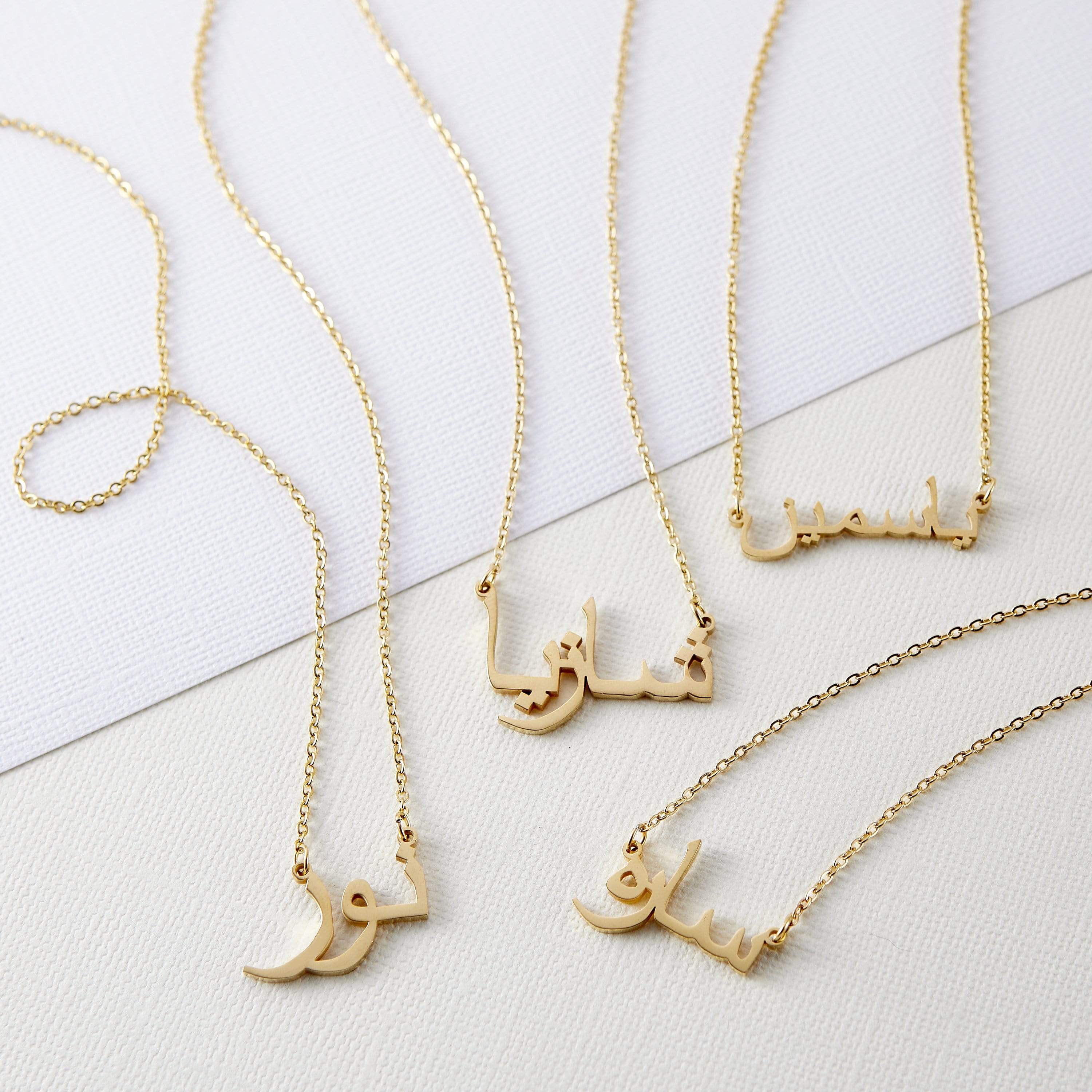 Four gold necklaces with pendants featuring different names written in Arabic script are arranged on a white rectangular tray. Each necklace has a delicate gold chain. The names on the pendants are distinct in their calligraphic style. The tray rests on a textured, light-colored surface.